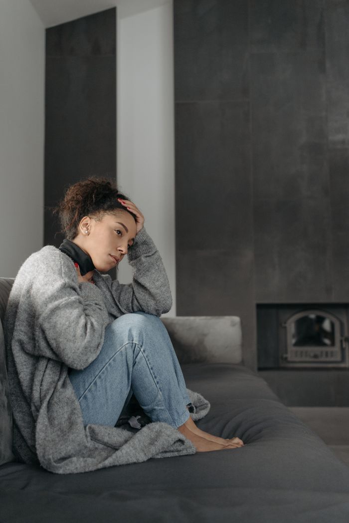 A woman sits on a sofa appearing unwell with a headache, wrapped in a cozy blanket in a modern room.